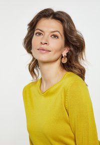 Pulover tricotat galben cu guler rotund. Modelul poartă cercei rotunzi, translucizi, care pun în evidență un ten neted și bucle ușoare.