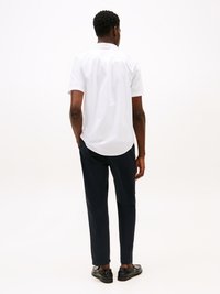 Uomo in piedi di spalle, con una camicia bianca a maniche corte, pantaloni scuri e mocassini neri su uno sfondo bianco uniforme.