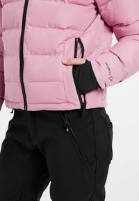 Rosa Daunenjacke mit horizontalen Nähten, schwarzen Reißverschlusstaschen und einem schwarzen Bündchendetail. Getragen mit schwarzen Hosen mit Reißverschlusstaschen.