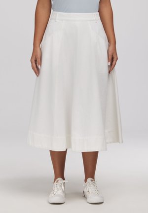 Kobieta ubrana w białą spódnicę midi z bocznymi kieszeniami, do której dobrano białe sznurowane trampki, stojąca na tle gładkiego tła.