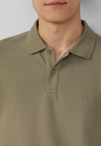 Olijfgroene poloshirt gemaakt van textuurstof. Beschikt over een omgeslagen kraag, twee knopen en een klein logo op de linkerkant van de borst.