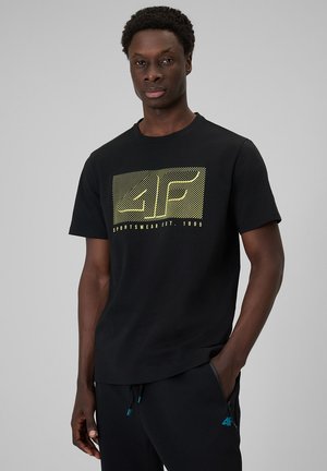 Mężczyzna ubrany w czarną koszulkę z żółtym logo 4F i napisem „Sportswear Est. 1995”, ręce w kieszeniach, stoi na tle jednolitego szarego tła.