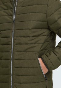 Nærbilde av en grønn polstret jakke med glidelås foran og sidelommer, som viser stoffets tekstur og quiltede mønster.
