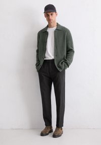 Groene geribde cardigan, witte t-shirt, donkere op maat gemaakte broek, bruine suède enkellaarsjes en een zwarte pet. Eenvoudig ontwerp met knopen en textuur.