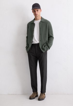 Green ribbed cardigan, white t-shirt, dark tailored trousers, brown suede ankle boots, and a black cap. Simple design with buttons and texture.