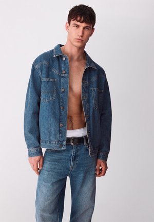 Man die een open blauwe spijkerjasje draagt, bijpassende jeans, zwarte riem en wit ondershirt zichtbaar boven de jeans, staande voor een effen achtergrond.