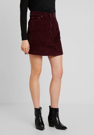 Femme portant une mini-jupe en velours côtelé bordeaux, un top à manches longues noir et des bottines noires, debout devant un fond gris uni.