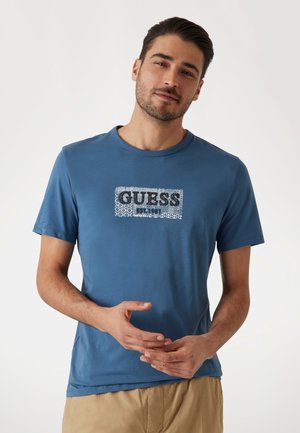 T-shirt en coton bleu avec un logo rectangulaire "GUESS" en noir, entouré d'une bordure à motifs. Manches courtes et design col rond.