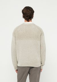 Άνδρας με καστανά σγουρά μαλλιά, φορώντας ένα υφασμένο μπεζ πουλόβερ και γκρι παντελόνι, στέκεται αντικρίζοντας ένα απλό λευκό φόντο.