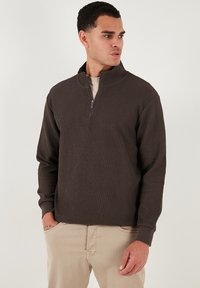 Pullover marrone a coste con colletto alto e chiusura a mezza zip. Maniche lunghe, tessuto testurizzato e vestibilità rilassata. Indossato sopra una camicia leggera.
