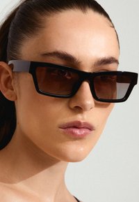 Ochelari de soare rectangulari negri cu blende maro, având rame groase și brațe elegante, așezați aproape de un ton al pielii deschis.
