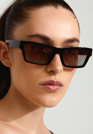 Black rectangular sunglasses with brown tinted lenses, featuring thick frames and sleek arms, positioned close to a light skin tone.