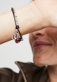 Pulsera de cuero con cuentas de plata, un charm morado y un acento en forma de corazón. El diseño es flexible y se enrolla alrededor de la muñeca.