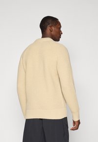 Pull en tricot couleur crème avec un motif texturé, col rond, poignets et taille côtelés. Présenté de dos sur un modèle.