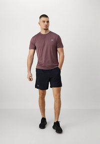 Νεαρός άνδρας στέκεται όρθιος, φορώντας μια μπορντό αθλητική μπλούζα, μαύρες αθλητικές βερμούδες και μαύρα αθλητικά παπούτσια, μπροστά σε απλό ανοιχτό γκρι φόντο.