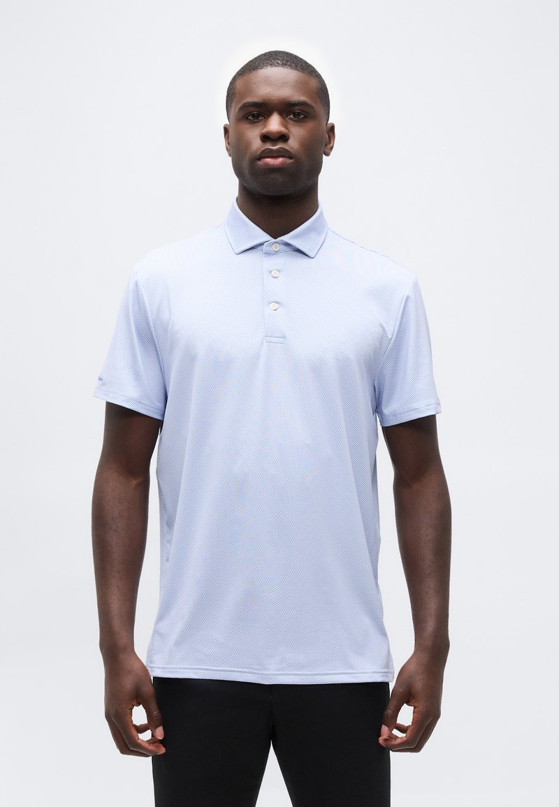 Άνδρας που φοράει ανοιχτό μπλε κον τομάνικο πόλο πουκάμισο με τρία κουμπιά και μαύρο παντελόνι, στέκεται μπροστά σε απλό λευκό φόντο.