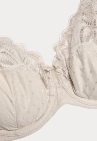 Beige kanten beha met gevoerde cups, een gestippelde textuur en een gegolfde kanten rand. Heeft een strikdetail in het midden en fijne stiknaden.