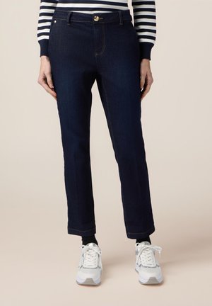 Pantaloni in denim blu scuro con vestibilità dritta, cinque tasche, chiusura con bottone dorato e cuciture a contrasto. Abbinati a sneakers bianche.