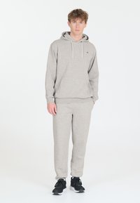 Hellgrauer Kapuzenpullover mit einer vorderen Tasche und Kordelzug, kombiniert mit passenden Jogginghosen. Schwarze Sneaker runden das Outfit ab.