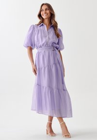 Lavender, tiered maxi dress with puff sleeves, button down front, belted waist, and sheer fabric layers. Paired with white ankle-strap sandals.