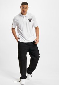 Vit polo skjorta med en logotyp, tillsammans med svarta cargobyxor. Outfiten inkluderar vita sneakers med en avslappnad passform och enkel design.