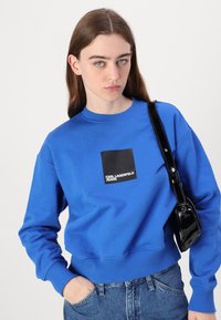 Felpa blu in cotone, con un logo quadrato nero che riporta la scritta "KARL LAGERFELD JEANS". Il design include polsini e orlo a coste.