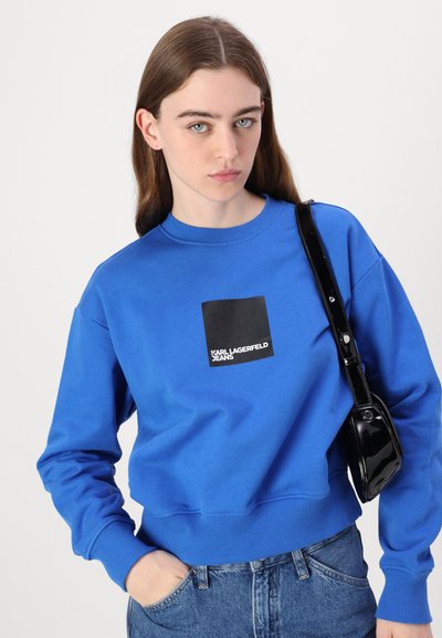 Sweatshirt bleu en coton, arborant un logo carré noir avec "KARL LAGERFELD JEANS". Le design comprend des poignets et un ourlet côtelés.