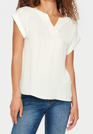 Vrouw met een wit blouse met korte mouwen en een V-hals, en blauwe spijkerbroek, staand tegen een effen witte achtergrond.