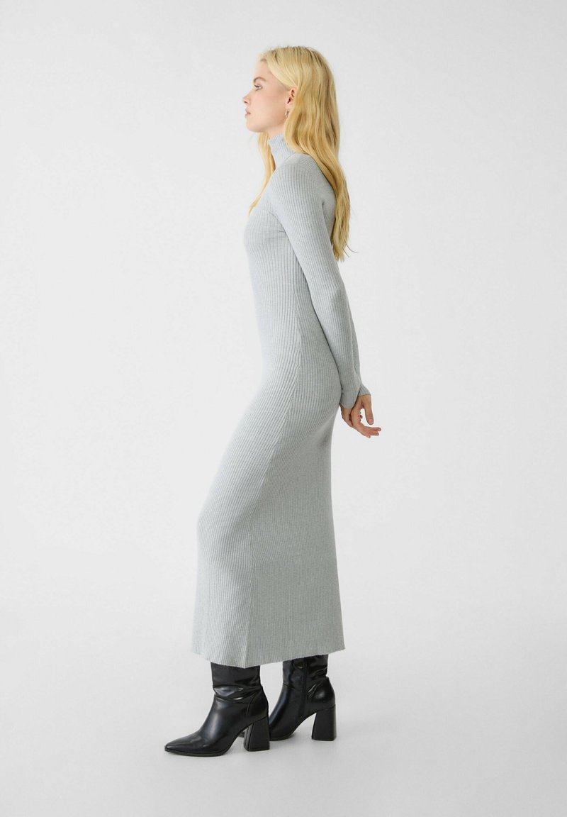 Vestido de punto acanalado gris claro con mangas largas y silueta ajustada, combinado con botas de tacón negro, mostrado de perfil.