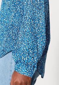 Blusa azul estampada com padrão de leopardo em preto e branco, com mangas longas e punhos franzidos. Textura leve e suave.