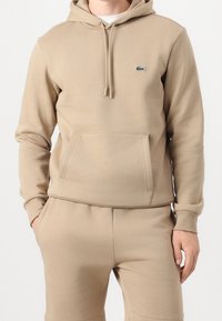 Homme portant un sweat à capuche beige et un pantalon assorti, les mains dans les poches, montrant le logo crocodile Lacoste sur la poitrine.