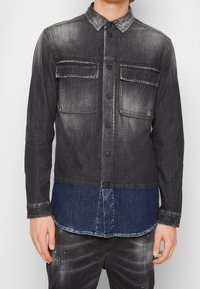 Camicia in denim con effetto ombre nero, dotata di due tasche sul petto, chiusura con bottoni e una parte inferiore blu a contrasto. Tessuto morbido e testurizzato.