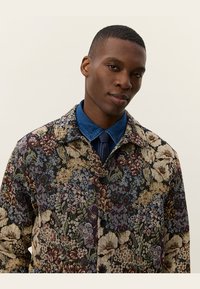 Floral-patterned jacket in multi-colors with a collared design. Features button closure, textured fabric, and a denim shirt underneath.