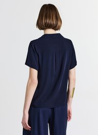 Chemise à manches courtes bleu marine avec une coupe décontractée, col et pinces au dos, conçue dans un tissu léger. Le modèle porte un bracelet texturé doré.