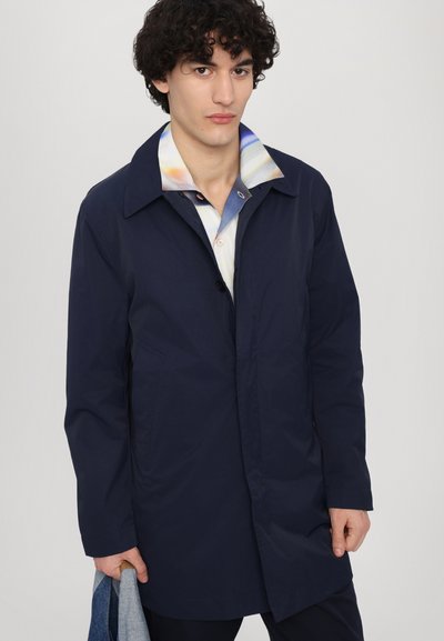 Imperméable bleu marine avec un col classique, boutonnage sur le devant et deux poches latérales ; porté par-dessus une chemise à motifs colorés avec une coupe décontractée.
