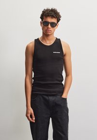 Jonge man met een zwarte zonnebril, een zwart mouwloos shirt met de tekst ""PEADOR"", een zwarte broek en een ketting als halsketting, staand tegen een effen muur.