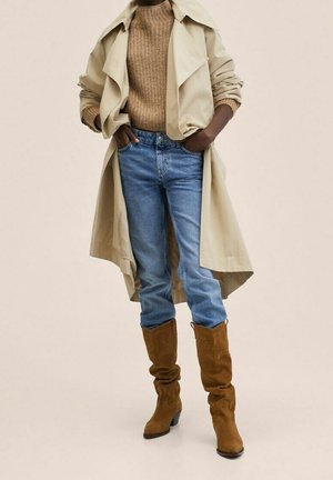 Henkilö, joka pukeutunut beigeen trenssitakkiin, ruskeaan neulepuseroon, sinisiin farkkuihin ja ruskeisiin mokkanahkaisiin keskipitkän varren saappaisiin, seisoo käsineenään taskut.