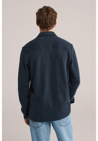 Muž v tmavomodrej košeli s dlhým rukávom a svetlomodrých džínsach, otočený chrbtom k jednoliatej béžovej stene.