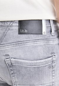 Grå denimjeans har en svart läderetikett med "l.t.b" på den högra bakfickan, och visar en urtvättad textur och snygg sömnad.