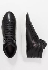 Baskets montantes en cuir noir avec une finition lisse, semelles en caoutchouc blanc et lacets gris. Présente des coutures ton sur ton et des accents sur les côtés.