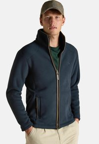 Tumesinise fleece-jakk, millel on kõrge krae, täispikk tõmblukk ja küljetaskud. Kantud rohelise särgi peal, mida täiendab mustriline pesapallikeep.