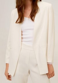 Blazer blanc avec un design structuré, légèrement à revers, et des poches avant, associé à un haut ajusté blanc. Texture lisse, coupe cintrée.
