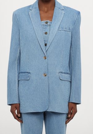 Vrouw draagt een lichtblauwe denim blazer met twee knopen, klepzakken en bijpassende high-waisted denim broek.