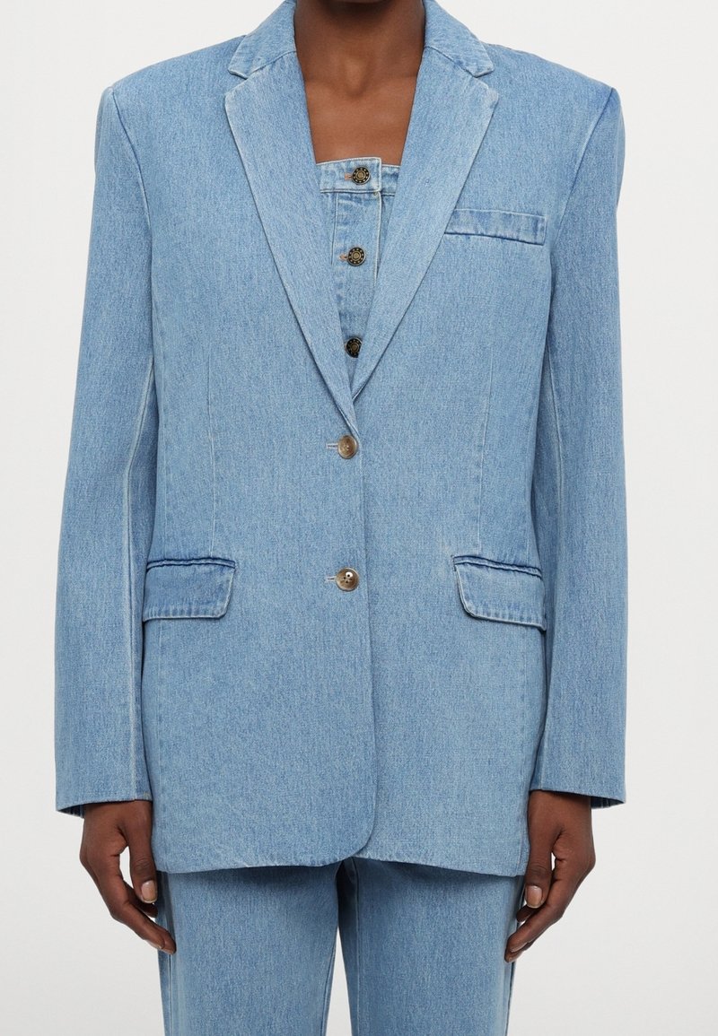 Femme portant un blazer en jean bleu clair avec deux boutons, des poches à rabat et un pantalon en jean assorti taille haute.