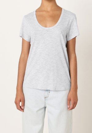 Persoon draagt een lichtgrijs T-shirt met korte mouwen en een ronde hals, en licht gewassen spijkerbroek, staat met ontspannen armen langs het lichaam.