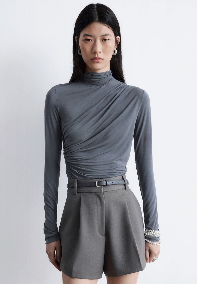 Haut gris à manches longues avec col montant et détail froncé à la taille, associé à un short gris taille haute, avec une ceinture large.