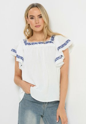 Frau mit langen blonden Haaren trägt eine weiße Bluse mit blauer Blumenstickerei an Ärmeln und Kragen, kombiniert mit hellblauen Jeans.