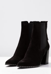 Botas de ante negras hasta el tobillo con tacones gruesos, paneles laterales elásticos y pequeñas tachuelas plateadas a lo largo de los bordes frontales.
