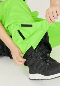Pantaloni impermeabili verde neon con polsini elastici neri e dettagli in Velcro, mostrati mentre vengono regolati su un stivale nero con suola bianca.
