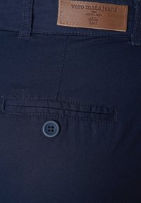 Jean en denim bleu marine avec une étiquette en cuir, un détail de poche avant et une fermeture à bouton unique. Texture lisse avec des accents cousus.